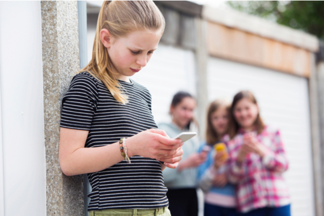 about-us-2 Young girl reading her phone with a puzzled expression. Girls behind her are huddled together with smiles watching her