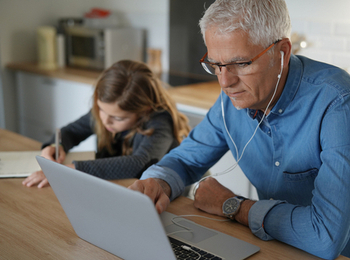 shutterstock_1674944041 1 Older man working on a laptop with earphones in. A young girl sits beside him writing on paper