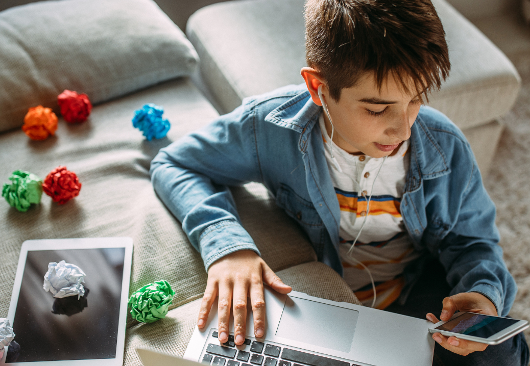 Child surrounded by balled up paper, a switched off tablet, laptop and scrolling a phone with ear phones in his ears